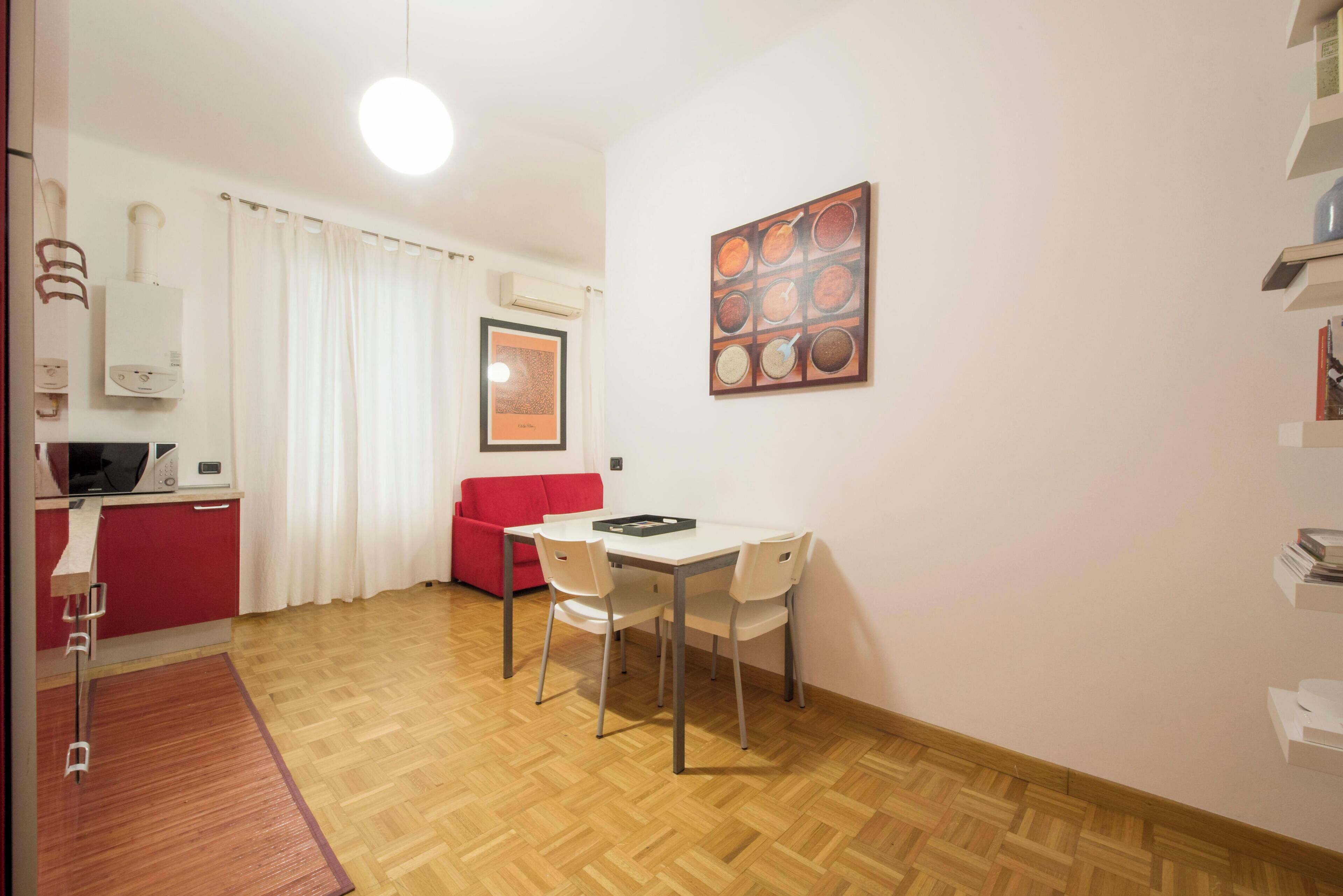 Ein kompakter Wohnraum mit Essbereich, einem roten Sofa, einer Mikrowelle und einem gerahmten Bild an der Wand.