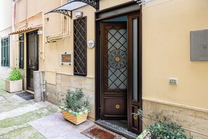 Um edifício com porta marrom, janela com grade de metal e pequena área de jardim com um cesto de plantas.