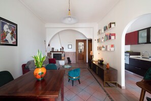 Ein modernes Wohnzimmer mit einem Holzespeisetisch, einem blauen Sessel und einer Pflanze. Es gibt eine Küchenzeile mit dunklen Schränken und einer weißen Arbeitsplatte.