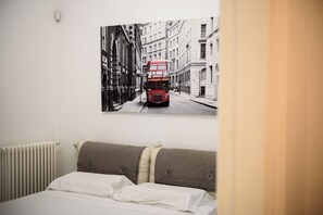 A hotel room with a double bed, a radiator, and a wall-mounted picture of a red double-decker bus on a city street. - Palazzo Dello Spagnolo Suite (Naples)