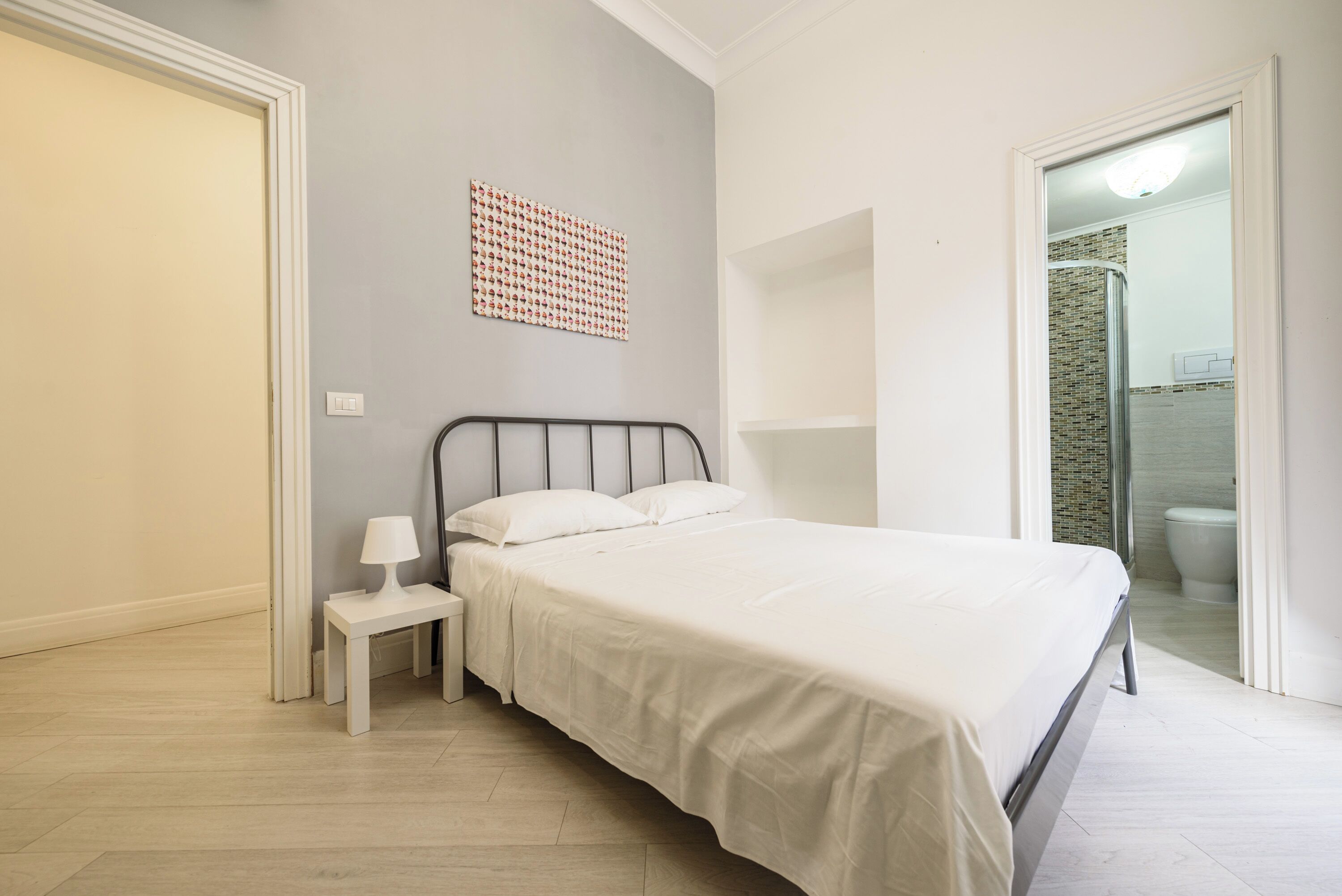 Quarto com cama, mesa de cabeceira, luz acoplada à parede e banheiro visível por uma porta aberta.