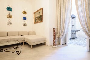 Een woonkamer met een beige bank, een schilderij aan de muur en uitzicht op een balkon door gordijnen.