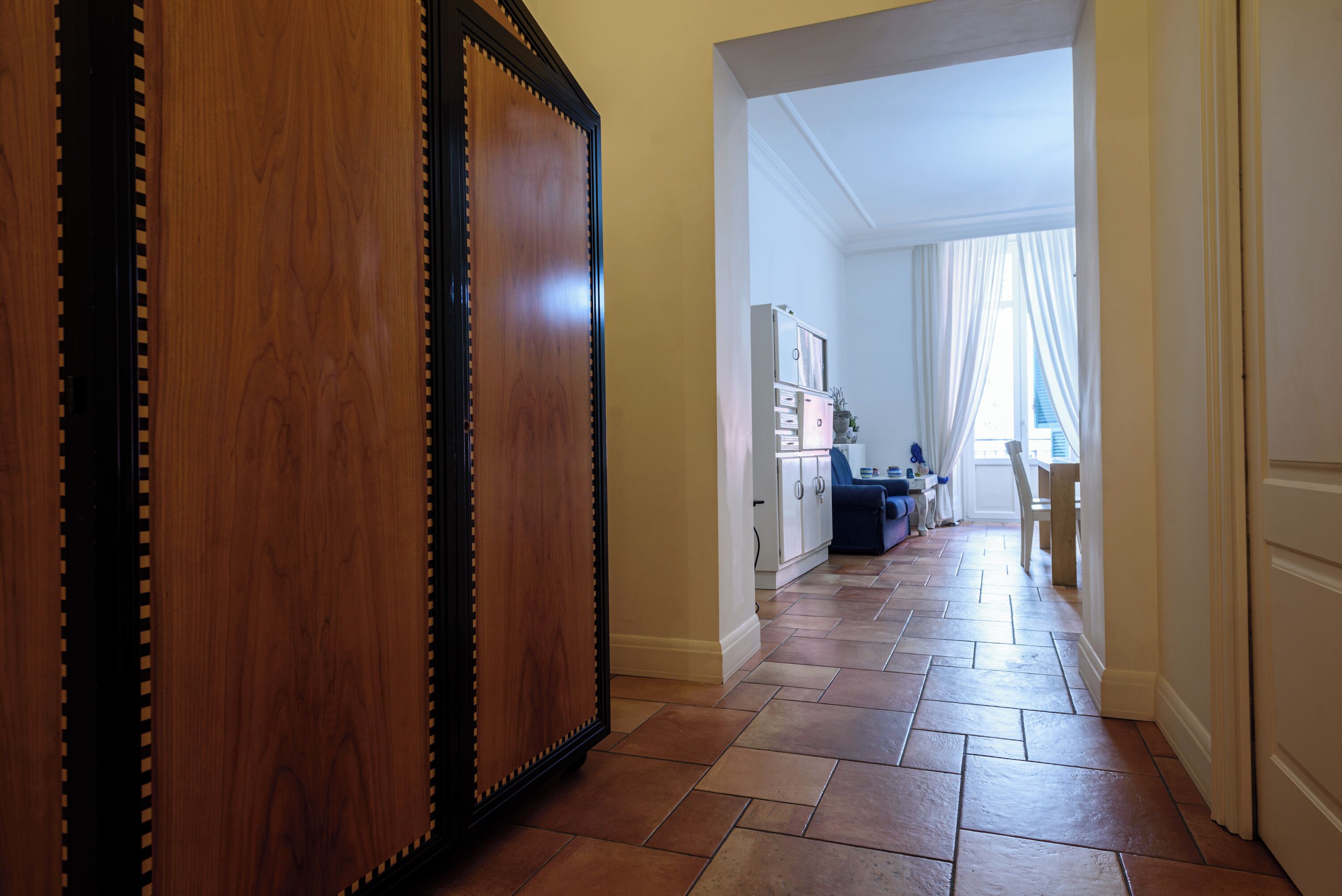 Un couloir avec une porte en bois, un sol carrelé et une vue sur une pièce dotée d’une fenêtre et de rideaux.