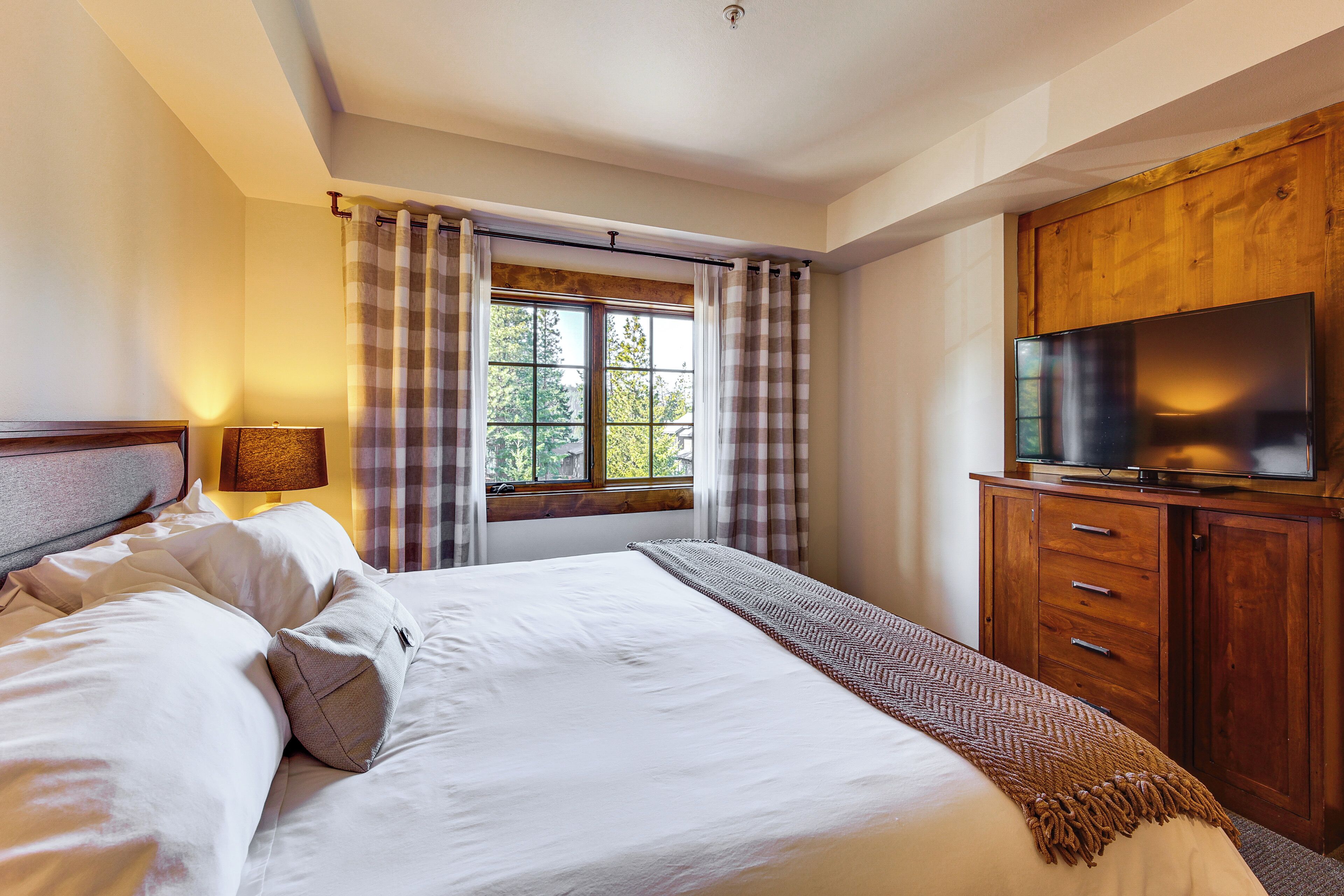 A hotel room with a bed, a TV on a wooden cabinet, a window with curtains, and a lamp.