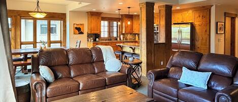 A spacious living room with leather sofas, a wooden coffee table, and a kitchen area with wooden cabinets and a refrigerator.
