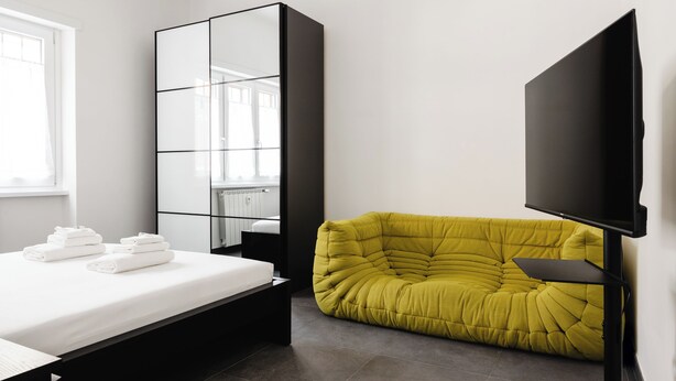 Quarto moderno com cama, espelho grande, sofá amarelo e televisão de tela plana.