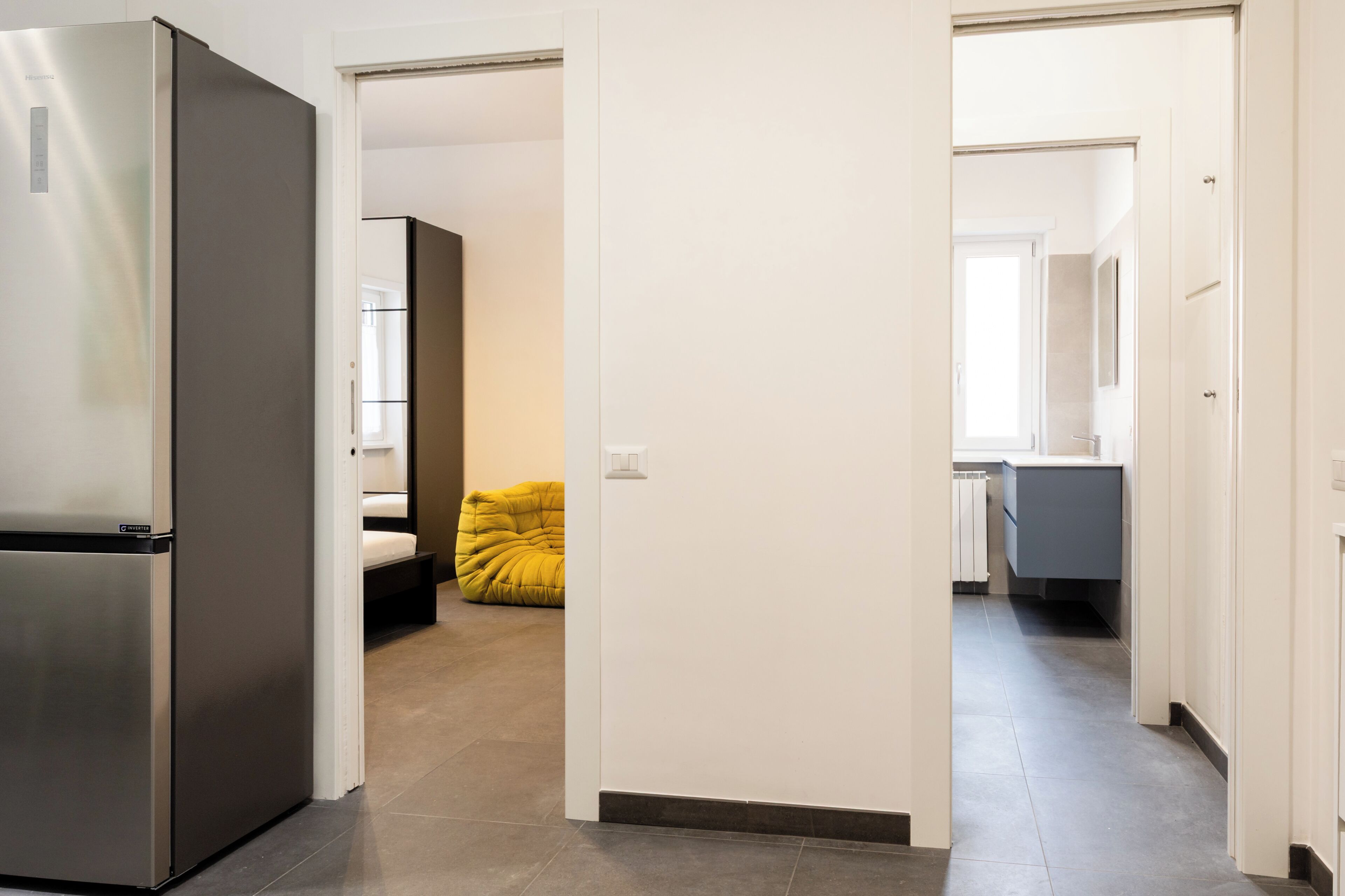 A modern kitchen with a built-in refrigerator, a dining area with a yellow chair, and a hallway leading to a bathroom.