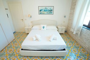 Un lit bien fait, avec du linge de lit blanc et deux serviettes enroulées, une table de chevet avec une lampe, une œuvre d’art encadrée au mur et un sol à motifs.
