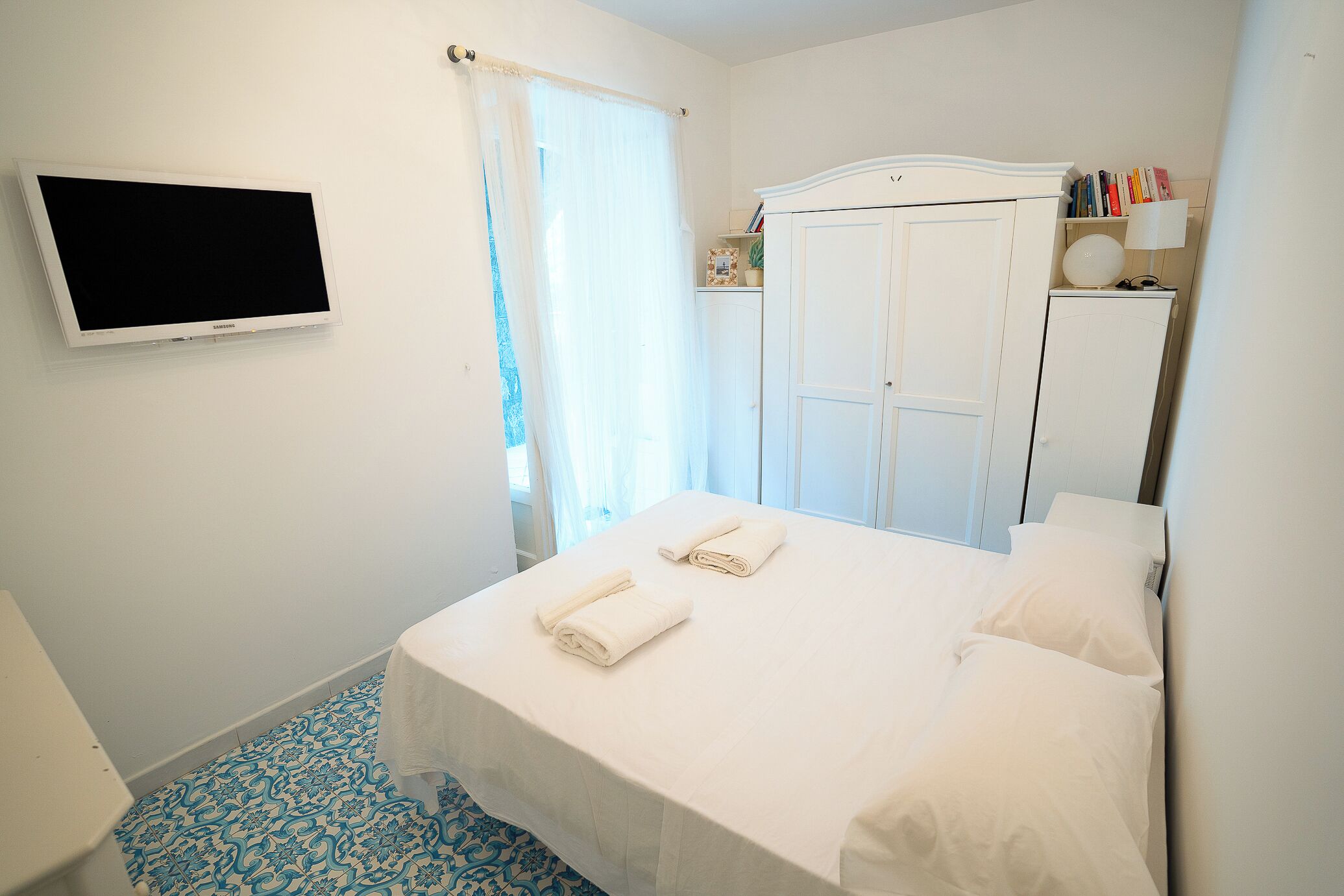 Una camera d'albergo con un letto, una TV a parete, un armadio e un comodino con una lampada.