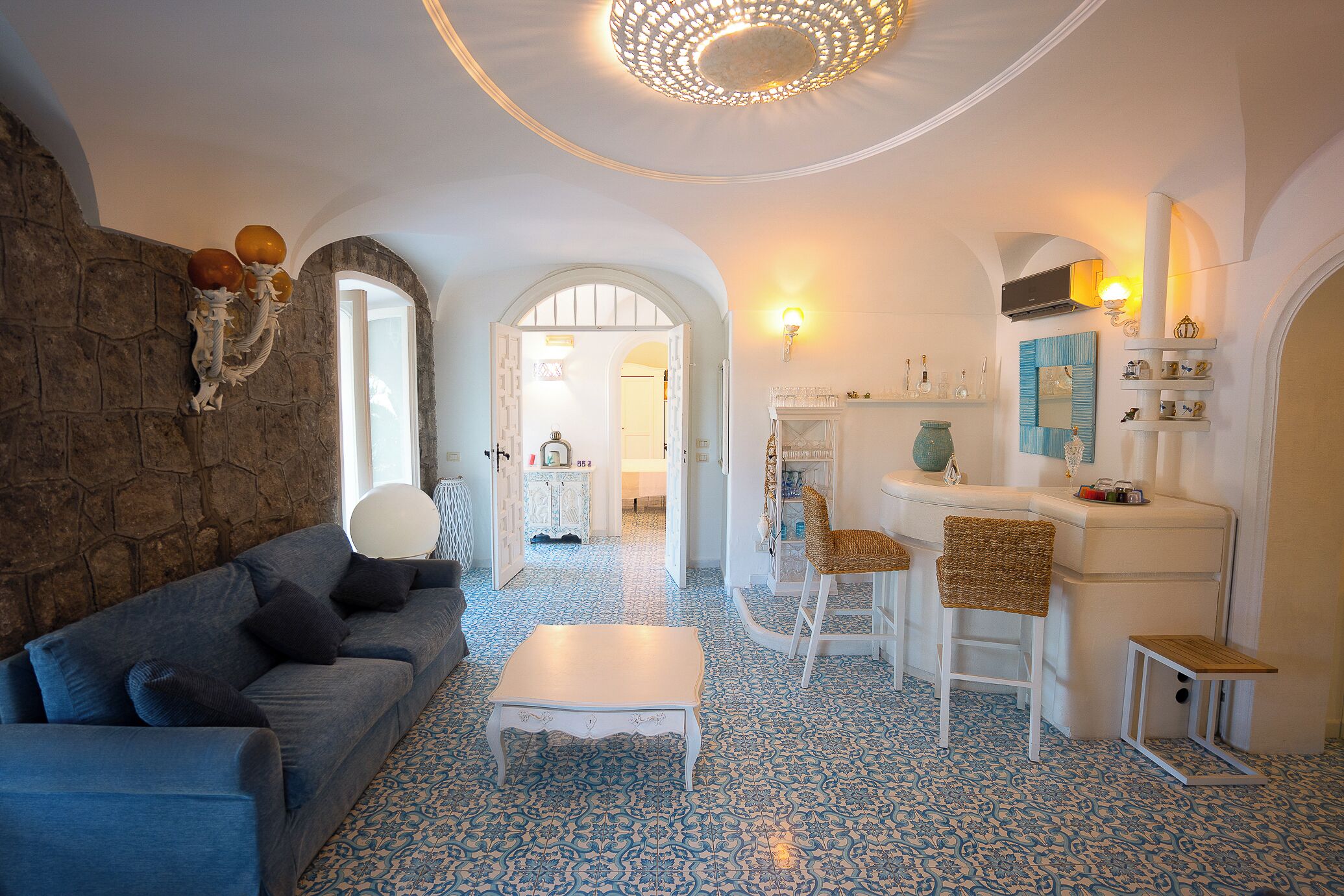 A living room with a stone wall, a blue patterned floor, a white coffee table, a grey sofa, and a wicker chair.