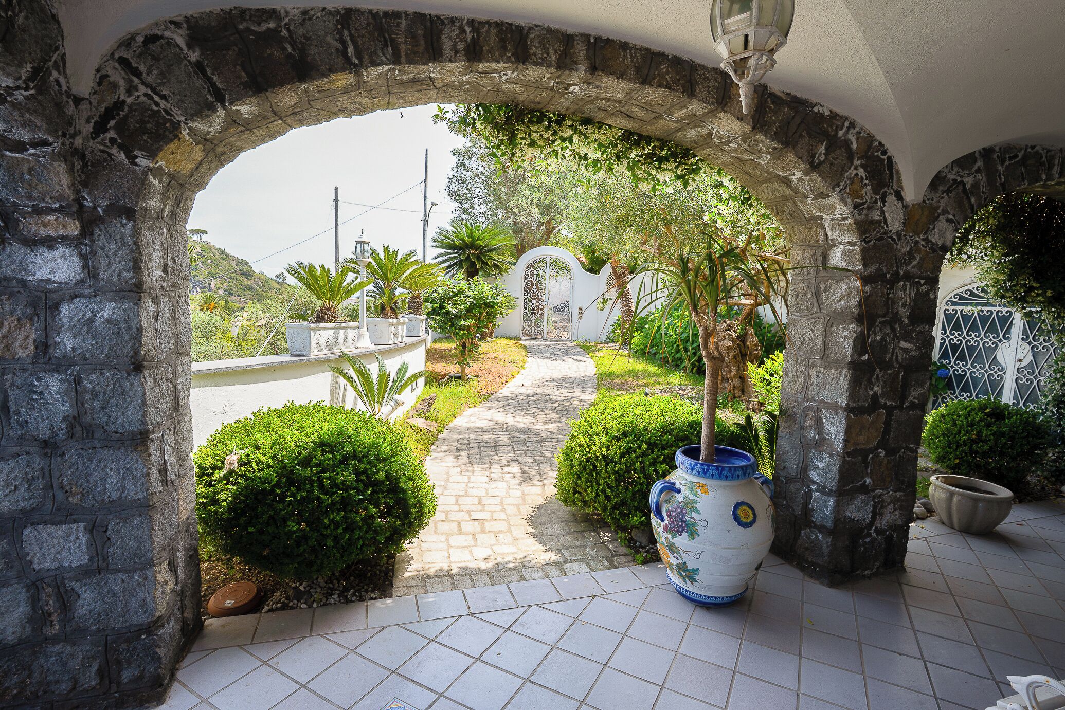 Un arco de piedra que da acceso a un jardín con un camino empedrado, plantas en macetas y un jarrón decorativo.