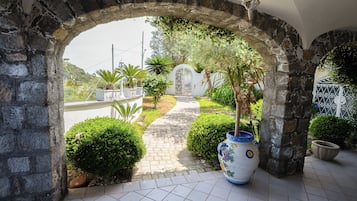 Un arco de piedra que da acceso a un jardín con un camino empedrado, plantas en macetas y un jarrón decorativo.