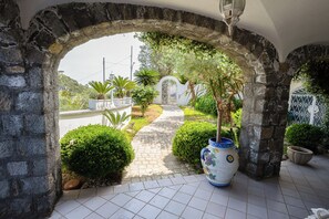 A stone archway leading to a garden with a tiled pathway, potted plants, and a decorative vase. - Villa Anna (Forio)