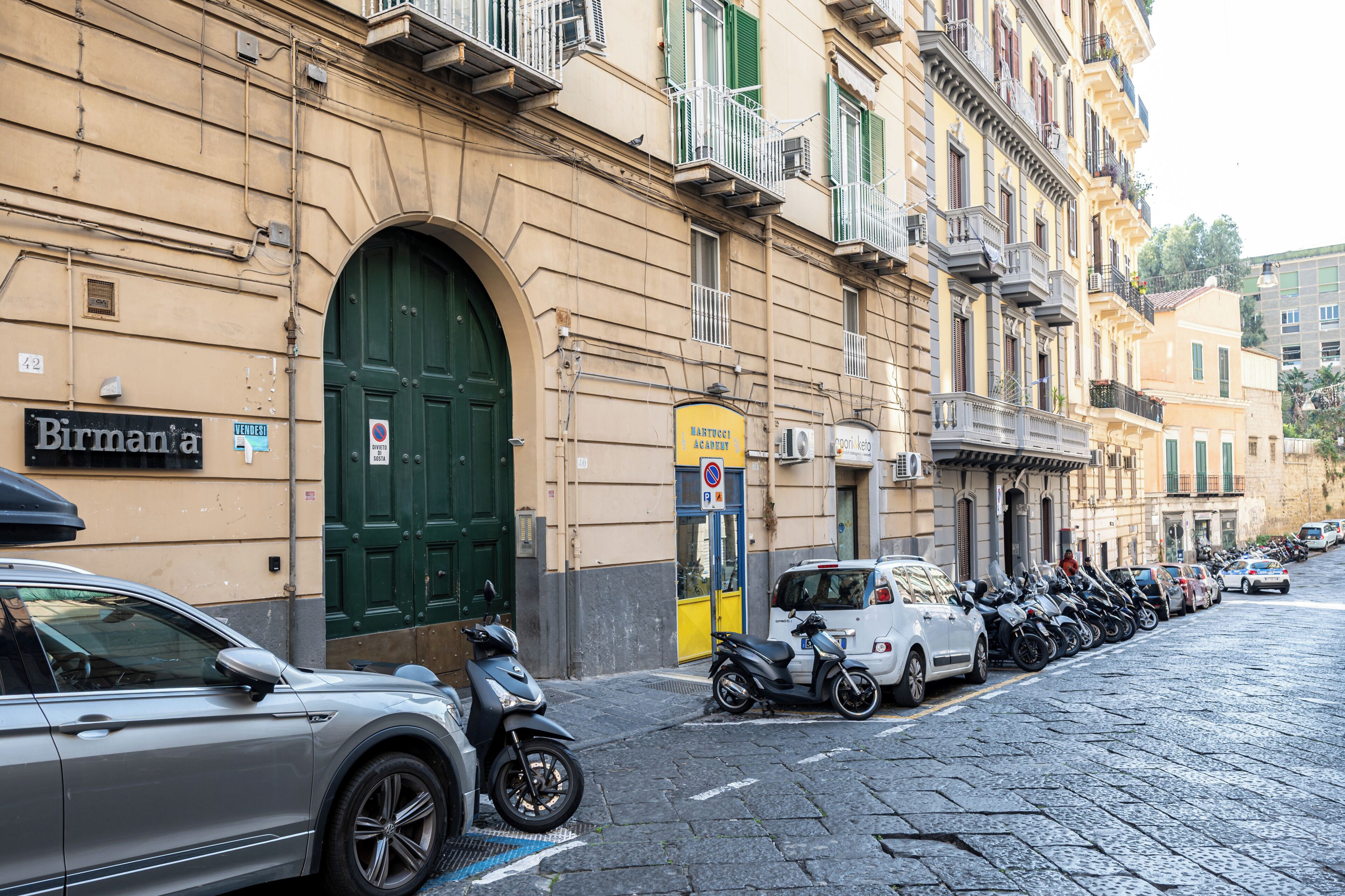 A street with parked cars and motorcycles, a building with a green door, and a sign that reads "Birmana".