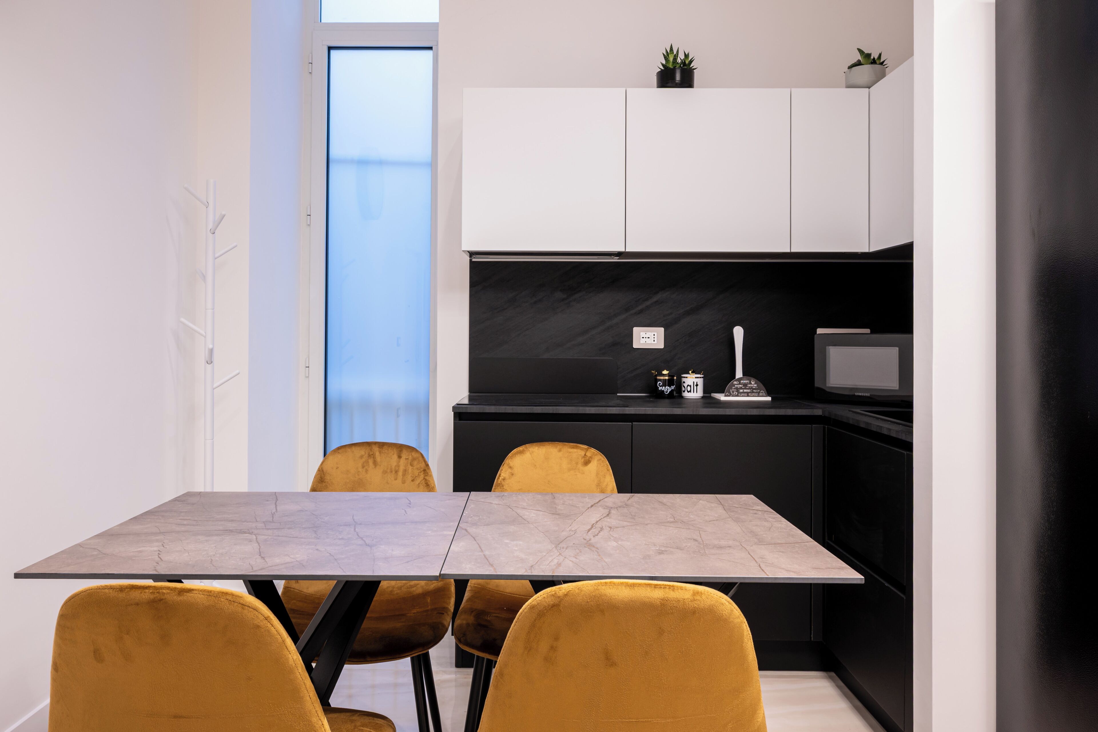 A modern kitchen with a black countertop, white cabinets, and a small potted plant on top. There is a dining table with chairs in front of the kitchen.