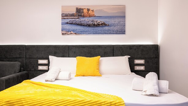 Habitación de hotel con cama, manta amarilla, almohadas blancas y un cuadro de una escena costera.