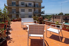 Terraza en la azotea con mobiliario exterior blanco, plantas en macetas y vistas a un edificio con balcones.
