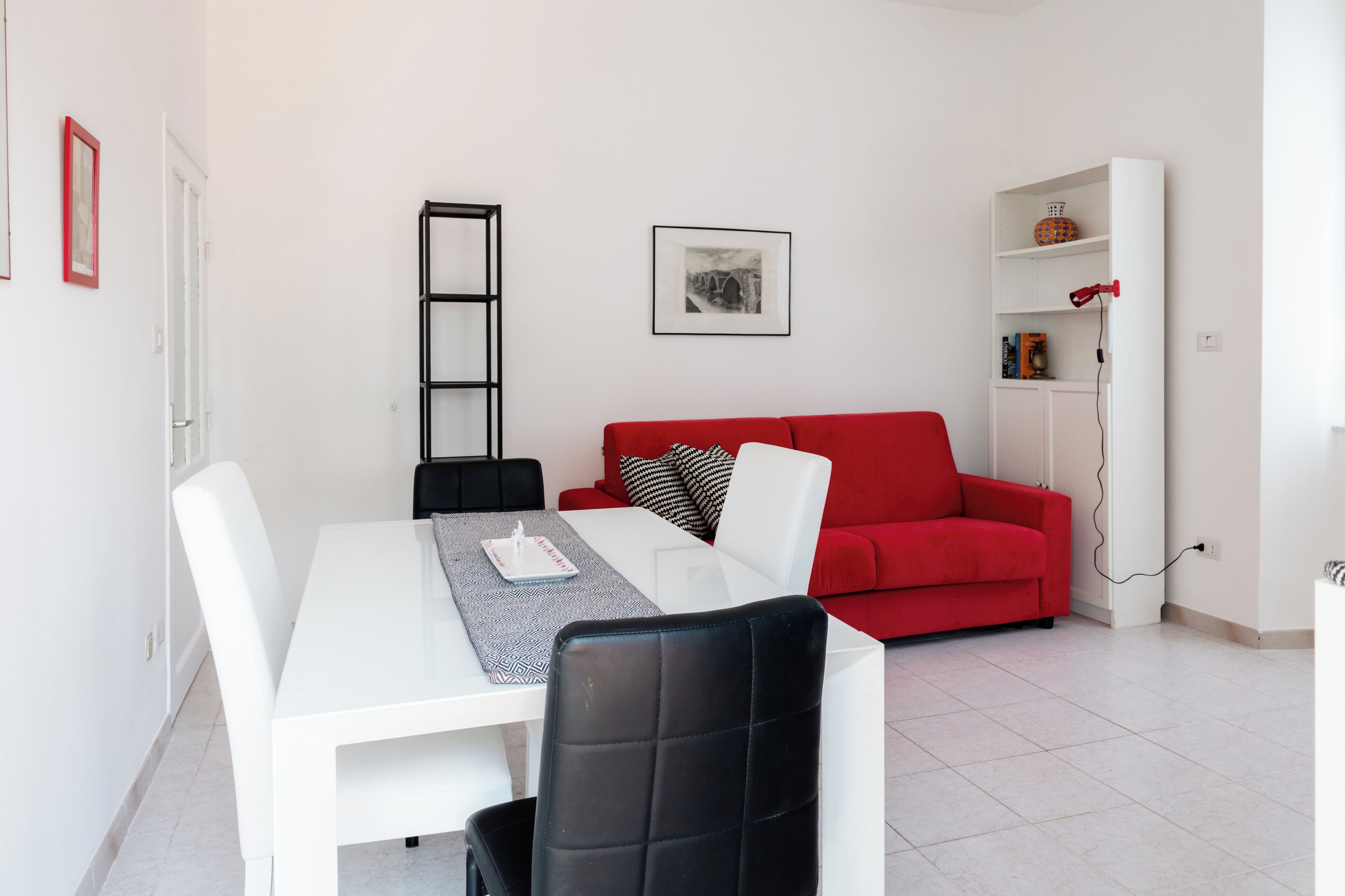 A modern living room with a white dining table, black chairs, a red sofa, and a black bookshelf.