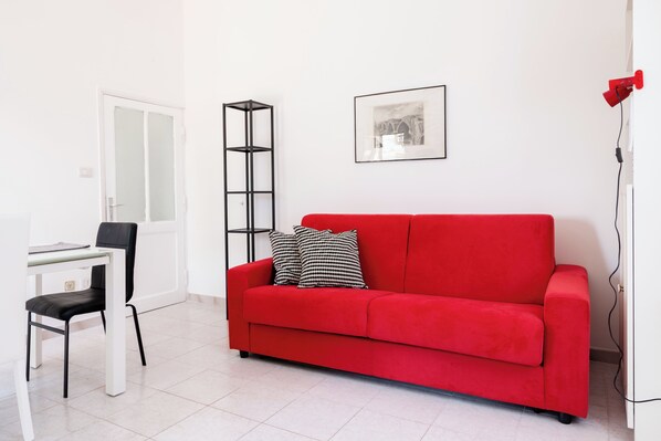 Un sofá rojo con almohadas de diseño blanco y negro, un escritorio blanco con una silla, un estante negro alto y un cuadro en la pared.