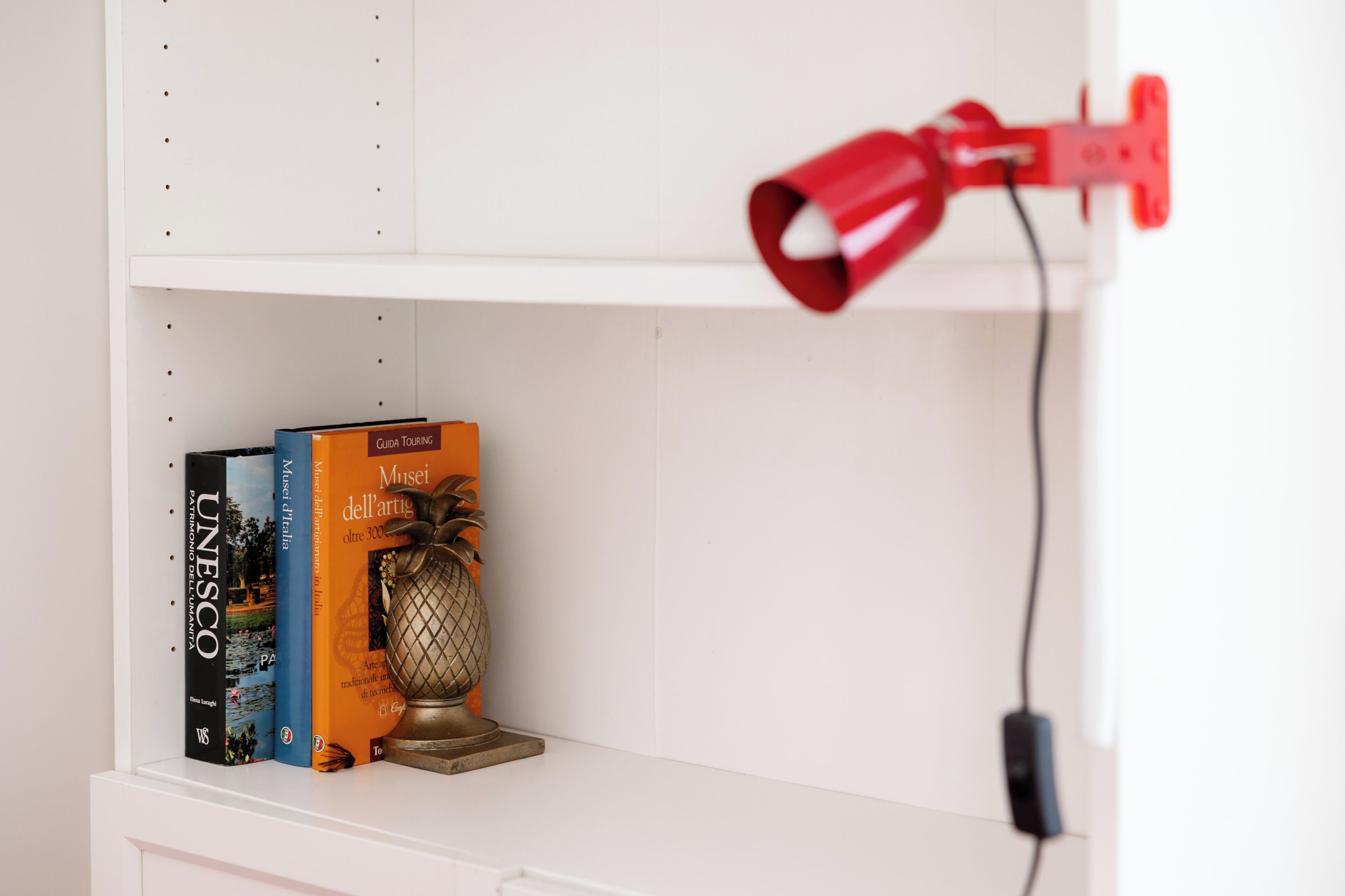 A white shelf with a red lamp, a pineapple, and three books titled 'UNESCO', 'Museo di Firenze', and 'Musei dell'arte'.