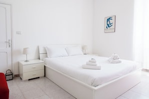 Un dormitorio blanco con cama, mesita de noche y una obra enmarcada en la pared.