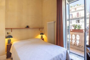 Ein Schlafzimmer mit einem Bett, zwei Nachttischlampen, einem Heizkörper und Blick auf einen Balkon mit Pflanzen und Gebäuden.