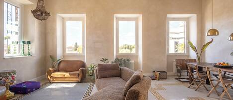 A spacious living room with a large sofa, a dining table with chairs, and multiple windows providing natural light.