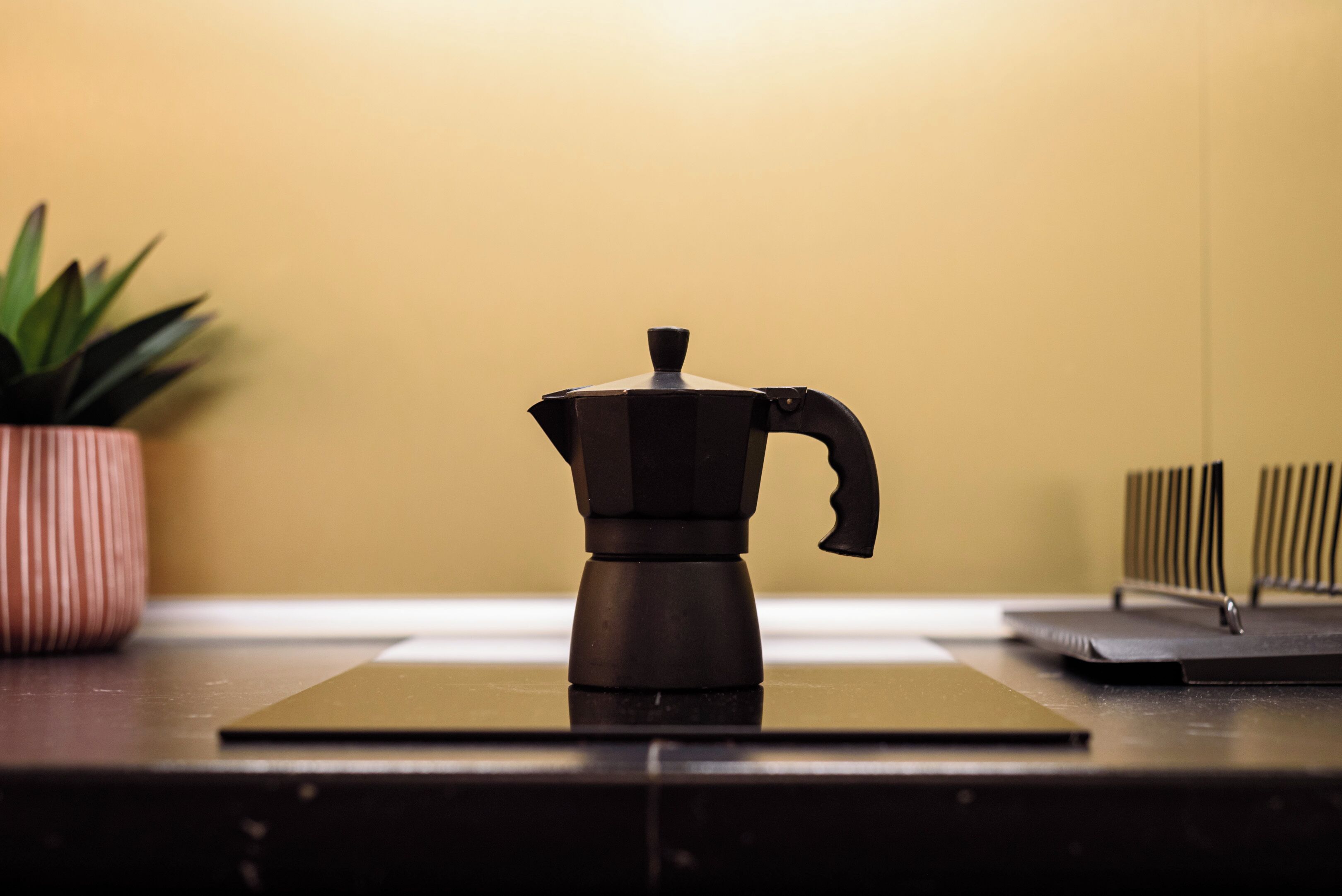 A coffee maker on a dark countertop with a potted plant and a radiator in the background.
