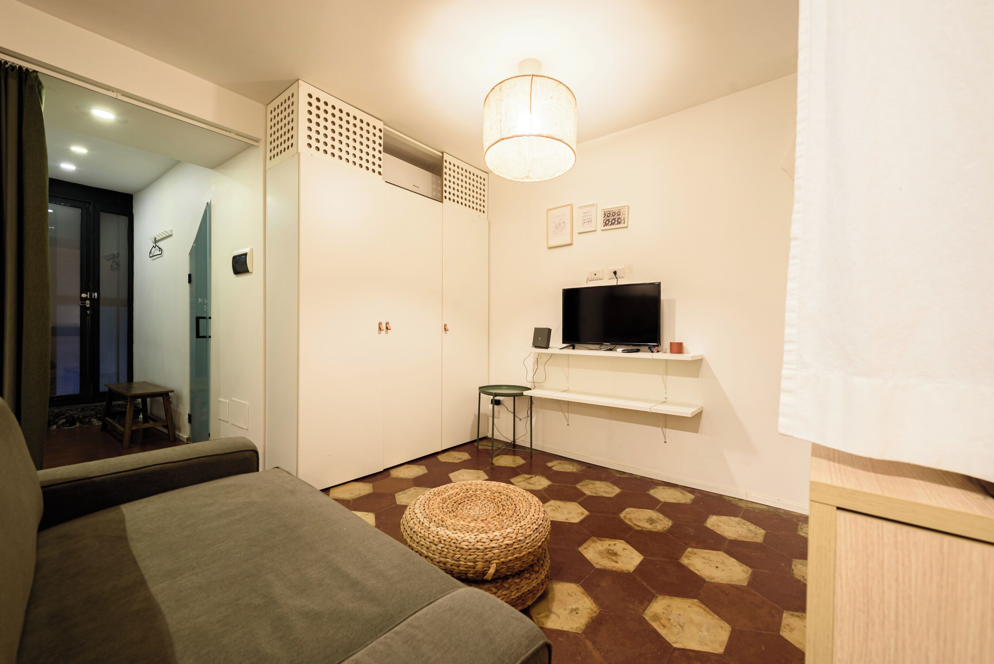 A small, well-lit room with a sofa, a television on a shelf, a woven ottoman, and a patterned floor.