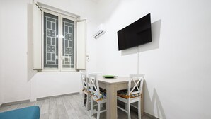 Une petite pièce propre, avec une table à manger, des chaises, un téléviseur fixé au mur et une fenêtre ornée d’une grille décorative.