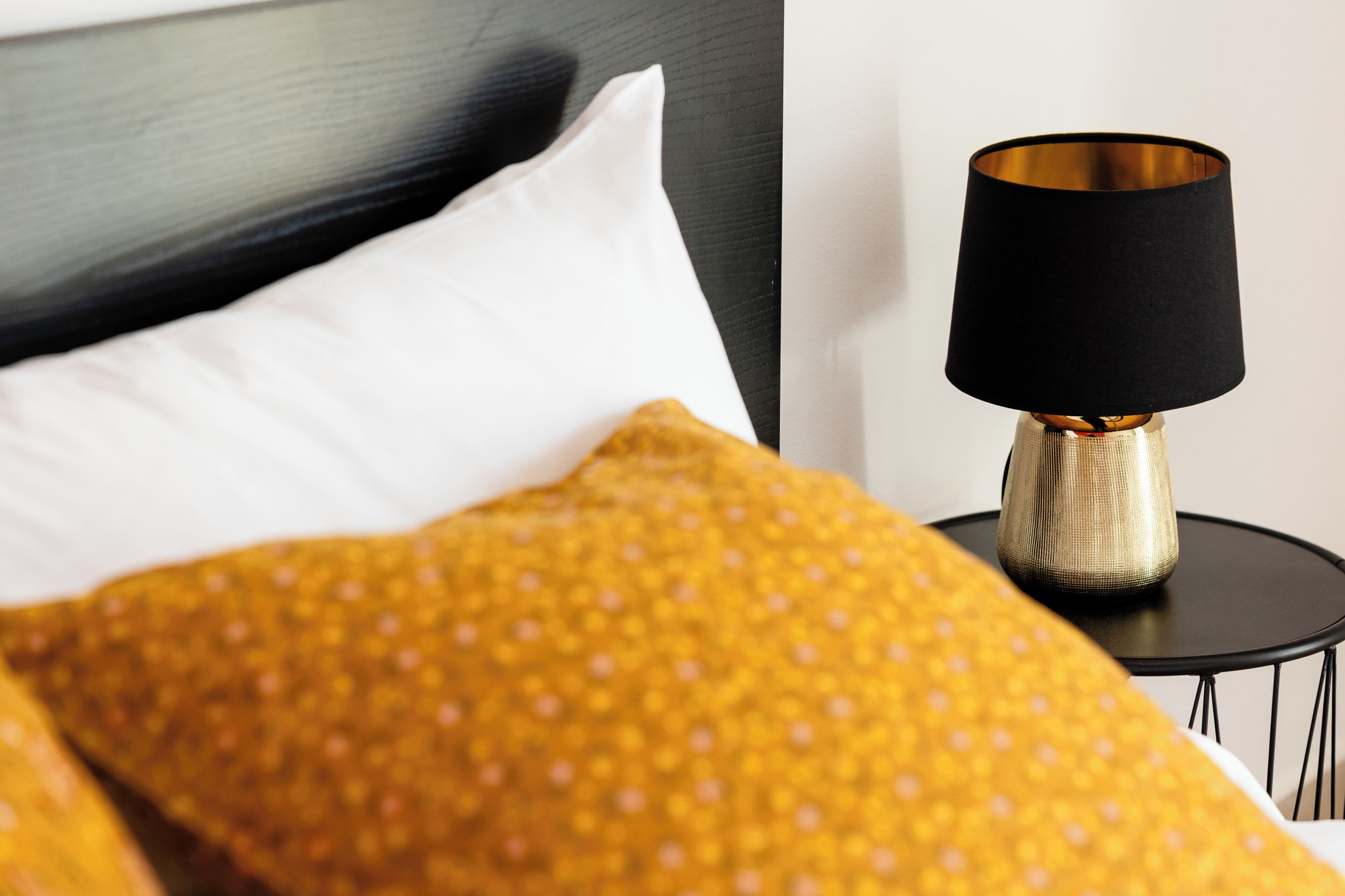 Ein Bett mit einem weißen Kopfkissen und einer gelb gepunkteten Decke, ein schwarzer Nachttisch mit einer Goldlampe und ein schwarzer Metallrahmen.
