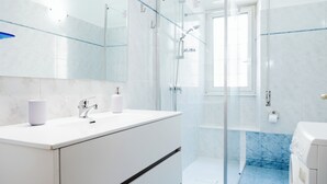 Een moderne badkamer met een glazen douchecabine, witte marmeren tegels en een witte wastafel met een spiegel erboven.