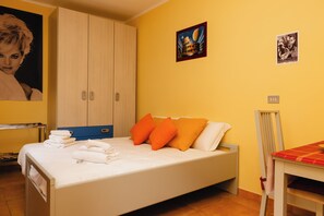 Habitación individual con una armario de madera, una mesita con mantel rojo y dos carteles en la pared.
