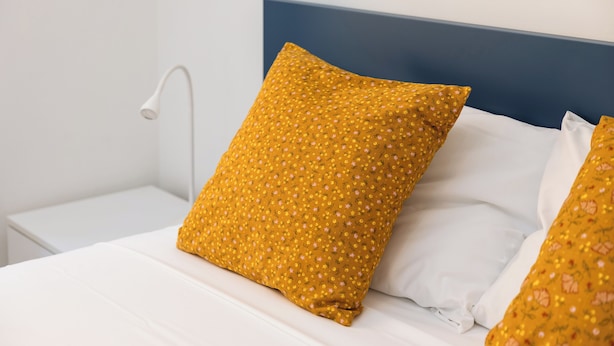 Ein ordentlich bezogenes Bett mit weißer Bettwäsche und einem senffarbenen Kissen mit Blumenmuster.