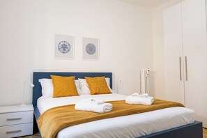 A neatly made bed with white linens, two mustard-colored pillows, and a brown throw blanket. There are two framed wall art pieces above the bed, a white nightstand with a lamp, and a white wardrobe with a handle. - Pandolfo Apartment (Rome)