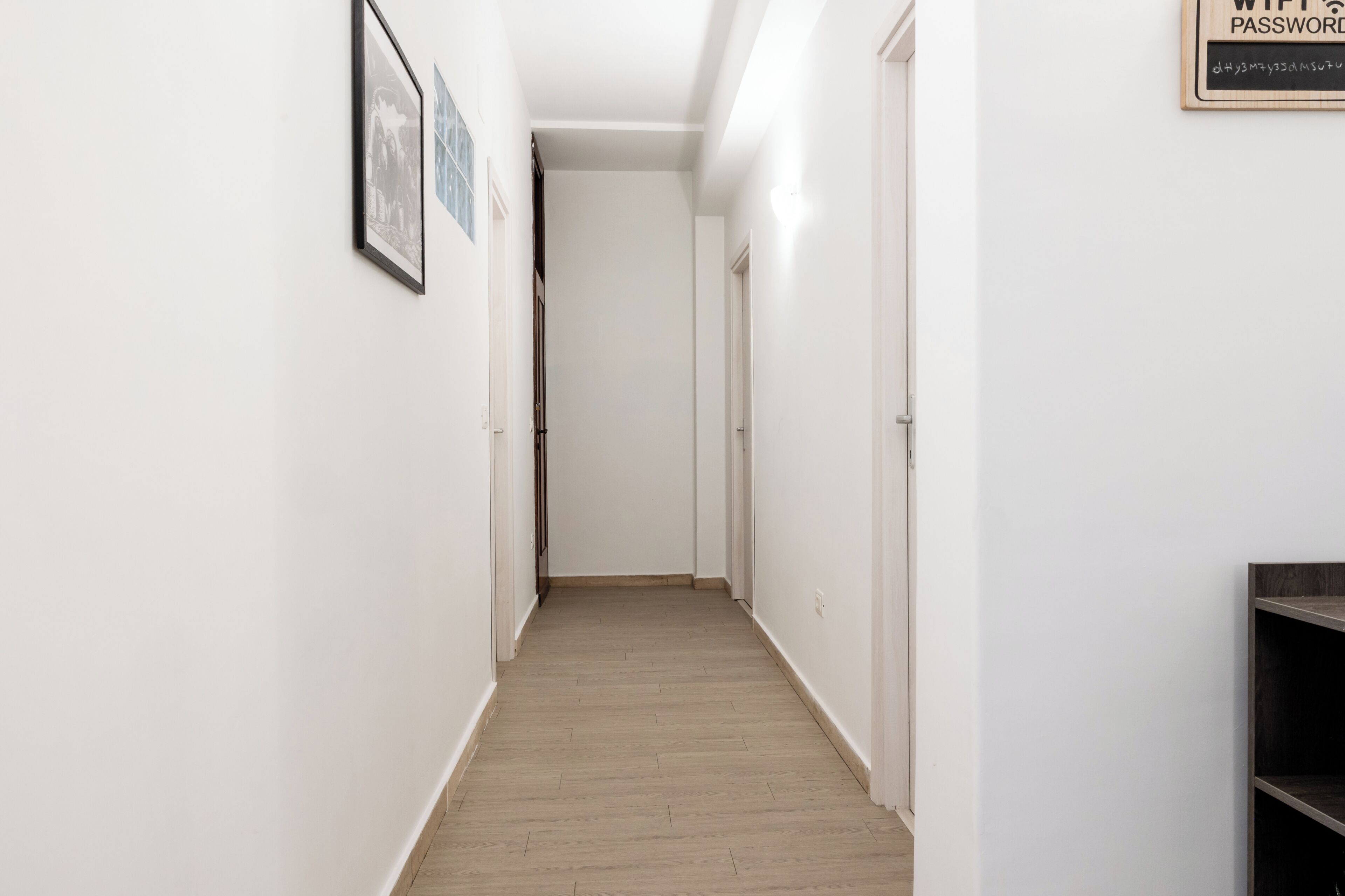 Un couloir étroit doté d’un plancher en bois et de murs blancs. Des tableaux encadrés ornent le mur de gauche et une enseigne est fixée au mur de droite.