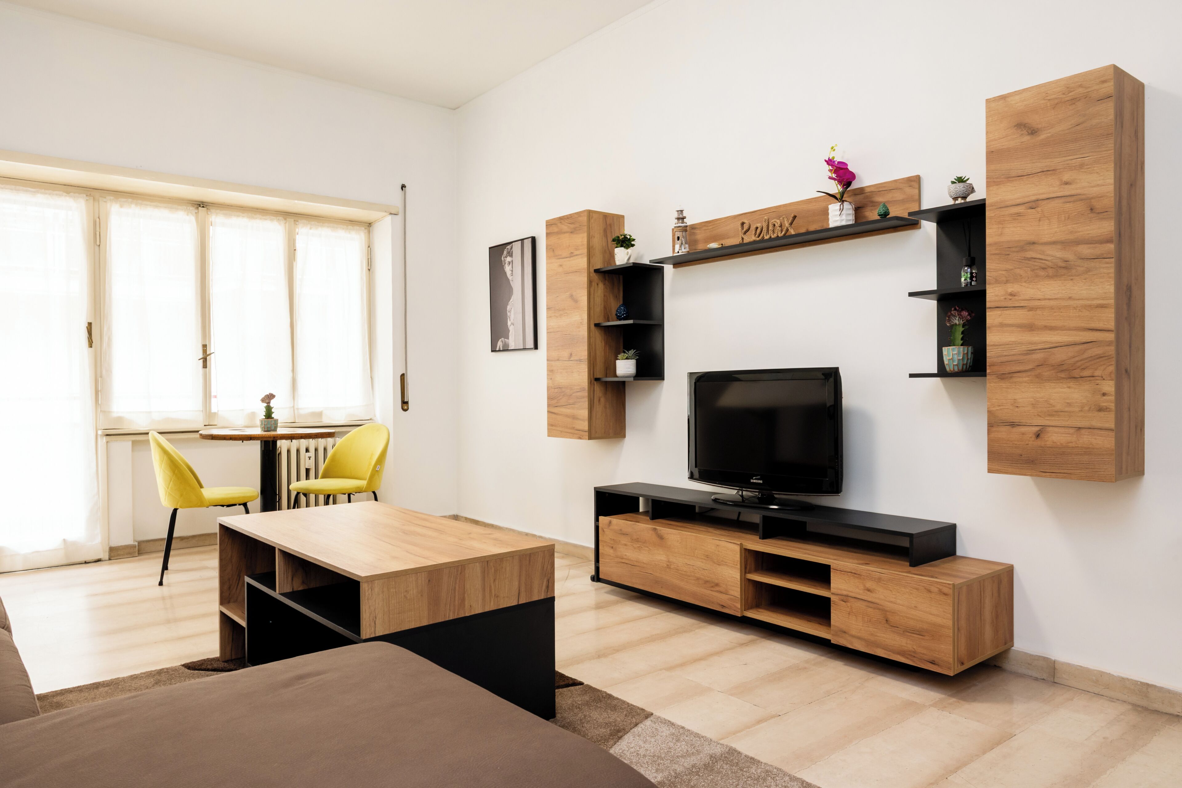 A modern living room with a wooden TV stand, a dining area with yellow chairs, and a large window.