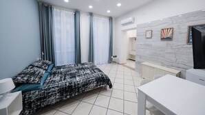Een slaapkamer met een bed, een bureau en een stoel. Er is een raam met gordijnen, een radiator en een wandairconditioner.