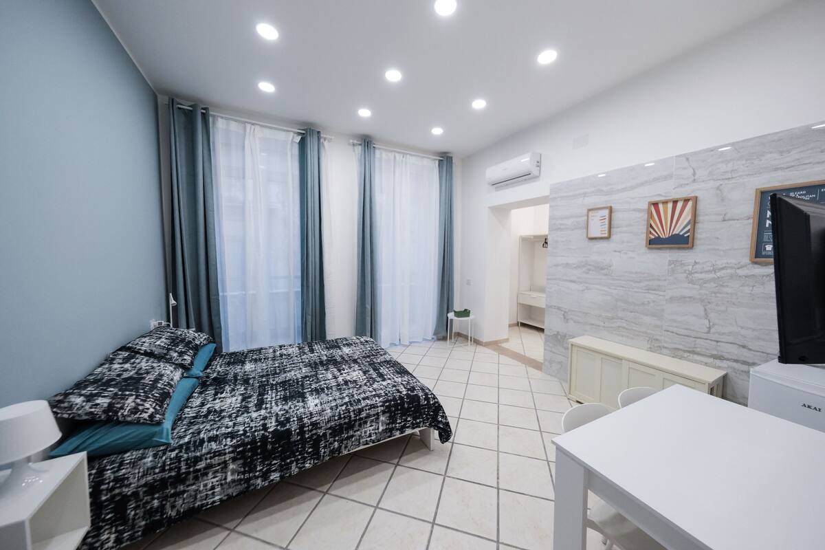 Ein Schlafzimmer mit einem Bett, einem Schreibtisch und einem Sessel. Es gibt ein Fenster mit Vorhängen, eine Heizung und eine Wandklimaanlage.