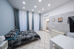 A bedroom with a bed, a desk, and a chair. There is a window with curtains, a radiator, and a wall-mounted air conditioner.