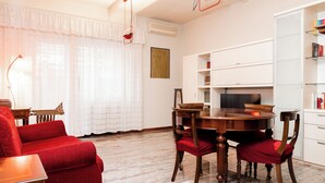 Ein Zimmer mit Esstisch, Stühlen, einem roten Sofa, einer Lampe und einem Bücherregal.