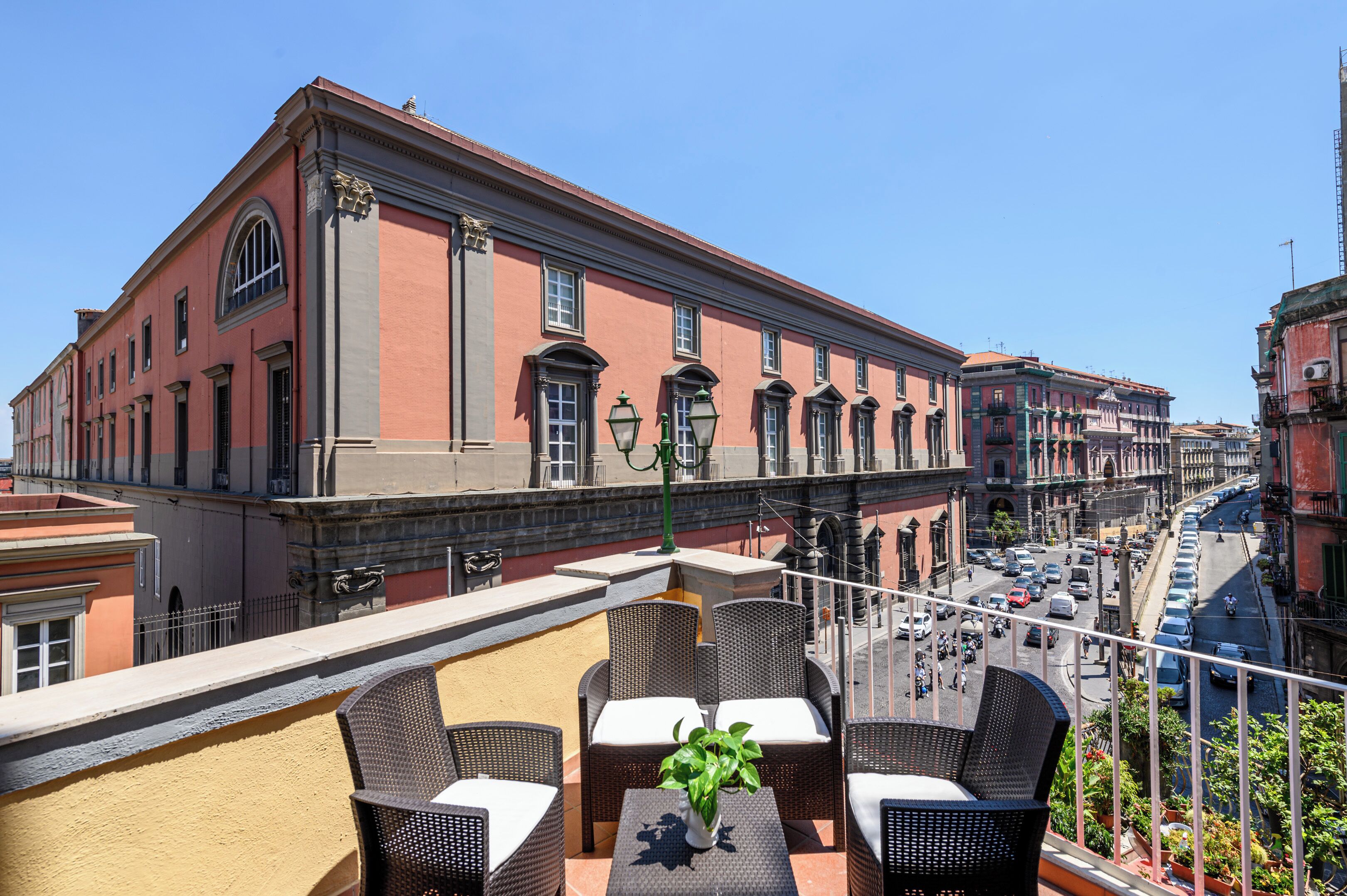 A terrace with wicker furniture and a potted plant, overlooking a street with historic buildings and parked cars.