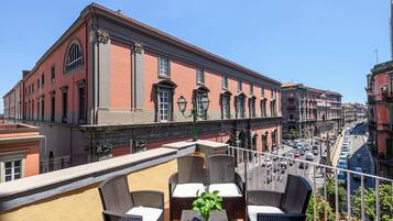 A terrace with wicker furniture and a potted plant, overlooking a street with historic buildings and parked cars.