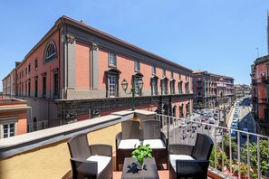 Une terrasse dotée de meubles en osier et agrémentée d’une plante en pot, donnant sur une rue bordée de bâtiments historiques et de voitures garées.