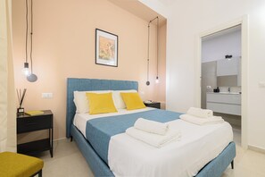 Un dormitorio ordenado con cama, mesitas de noche, un banco y un baño visible a través de una puerta abierta.