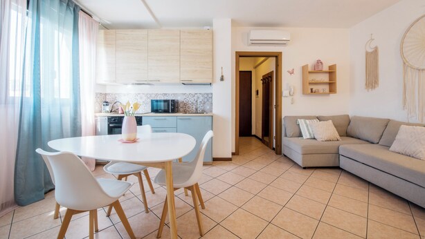 Una cocina moderna con armarios de madera clara, una mesa de comedor blanca con cuatro sillas y un sofá gris en la sala.