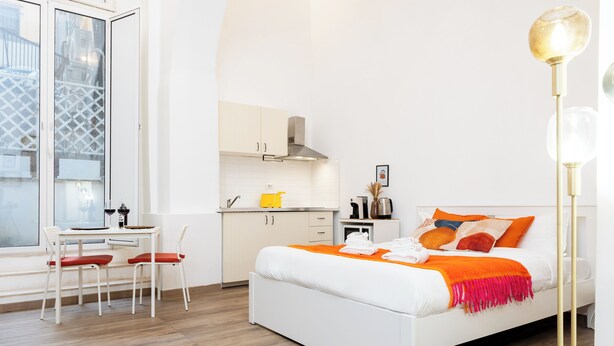 A modern bedroom with a white bed, orange throw, and a dining area with a yellow chair. - Archetto by the Vatican (Rome)