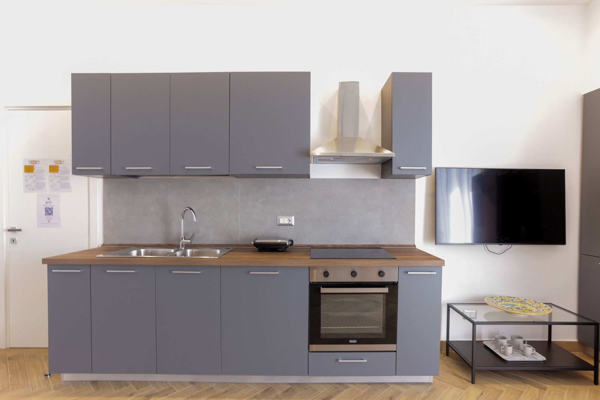 A modern kitchen with grey cabinets, a stainless steel sink, a built-in oven, and a flat-screen TV mounted on the wall.