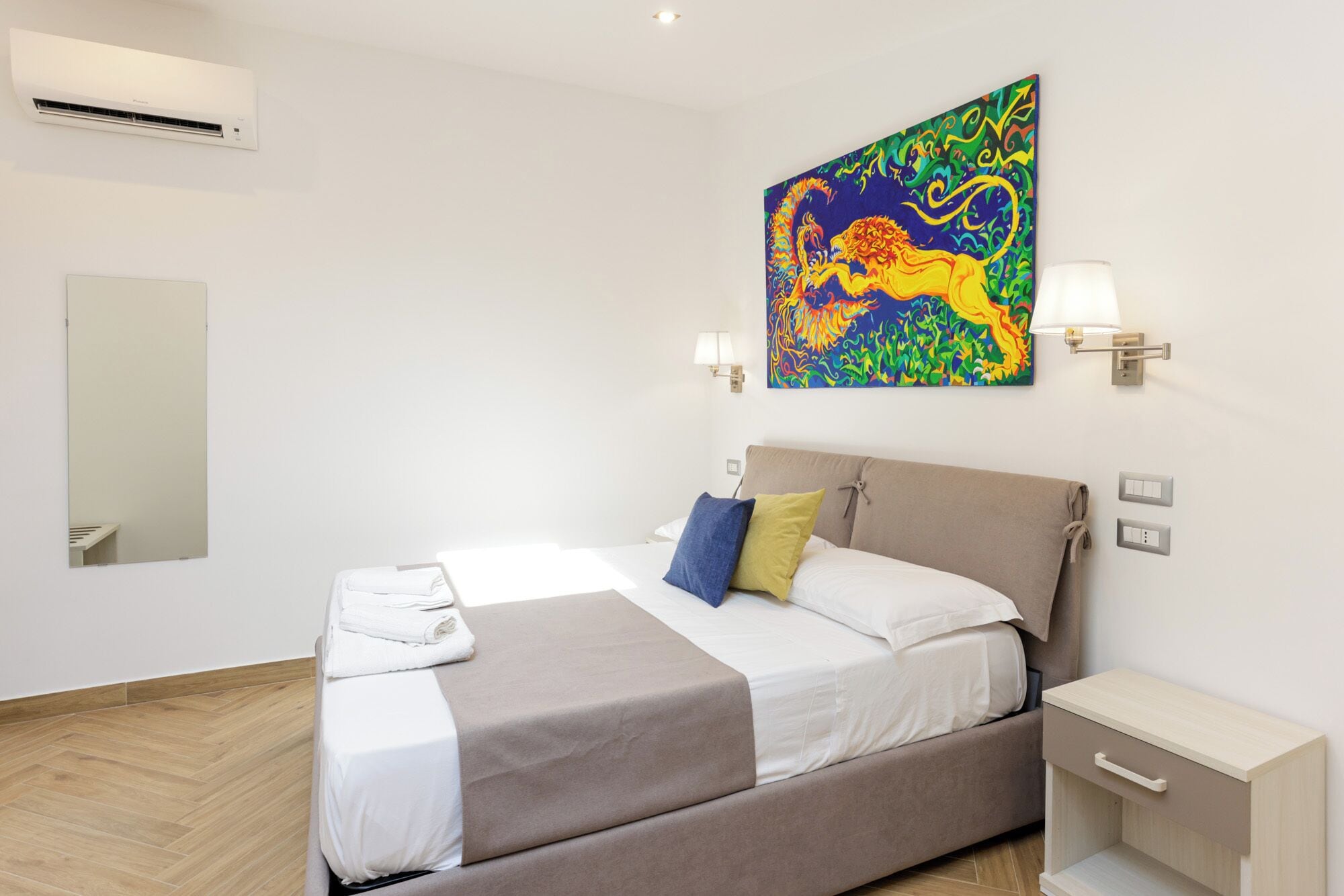 A modern hotel room with a bed, a nightstand, a lamp, and a colorful abstract painting on the wall.