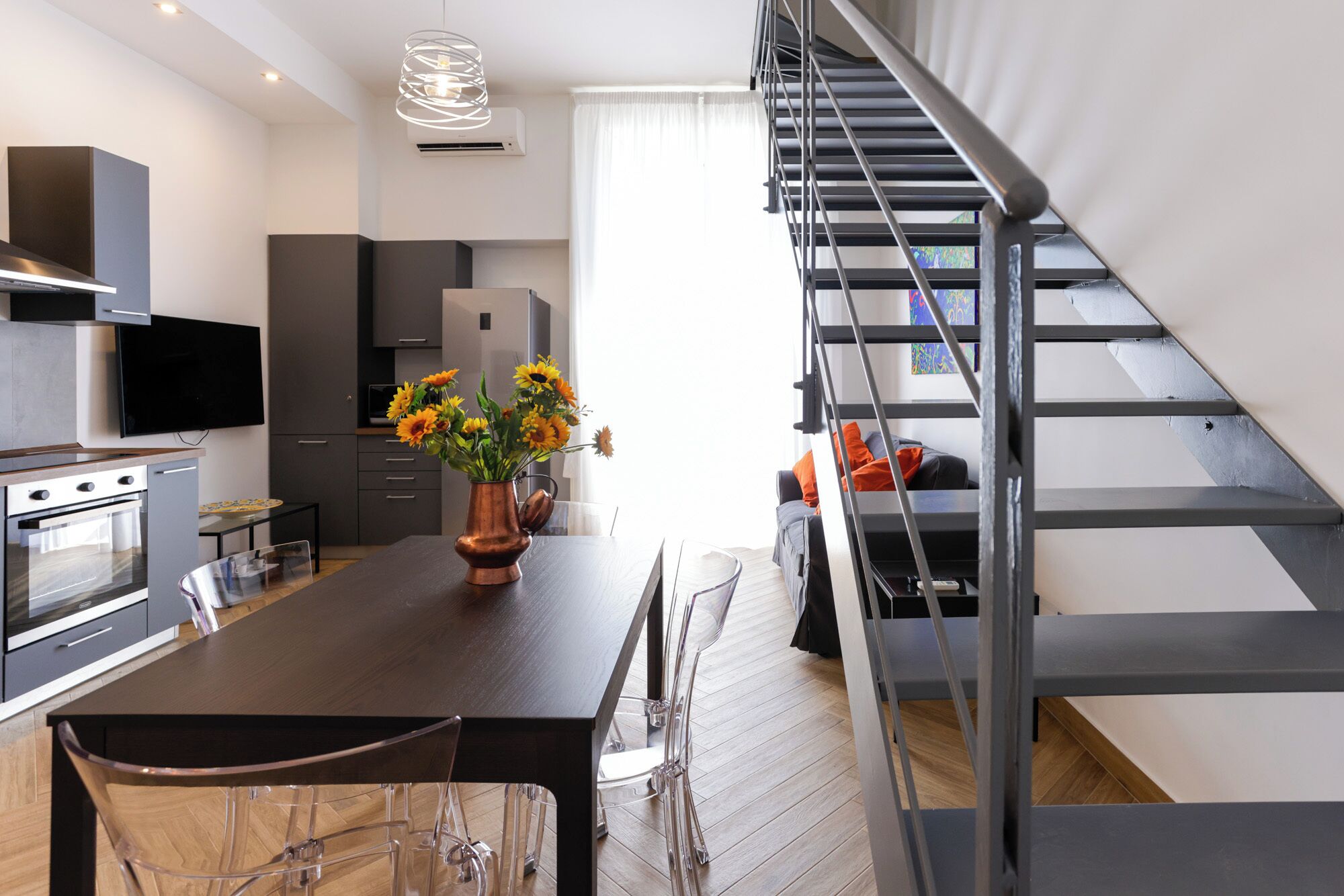 Eine moderne Küche mit einer zentralen Kochinsel, einem Essbereich mit transparenten Stühlen, einer Treppe und einem wandmontierten Flachbildfernseher.